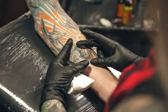 A tattoo artist working on a black and white greyscale tattoo of Anubis on a man's forearm.  An Anubis tattoo meaning differs according to what aspect of Anubis is significant to the person wearing the tattoo.