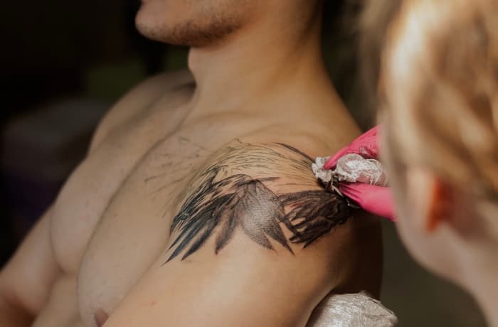 Tattoo artist creating wings and a bird's head on a man's shoulder