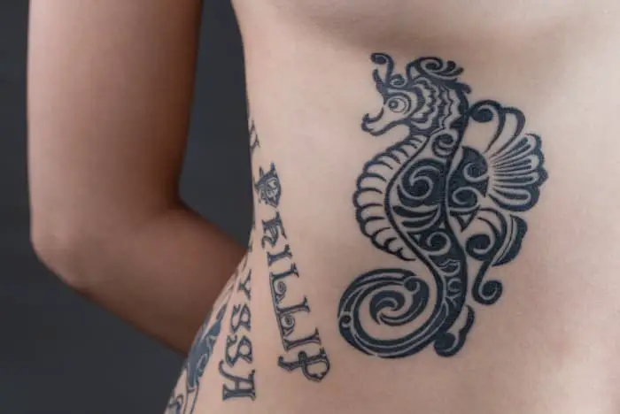 Black ink tattoos of names along with side of a woman's torso, along with a seahorse tattoo next to the names.