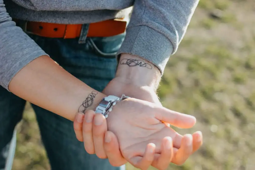 Couple showing their wrists where a tattoo design for couples with their initials interwoven is on each wrist.