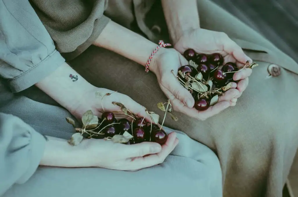 Two women holding handfuls of cherries, one of them with a small, delicate anchor and heart tattoo near her inner wrist.