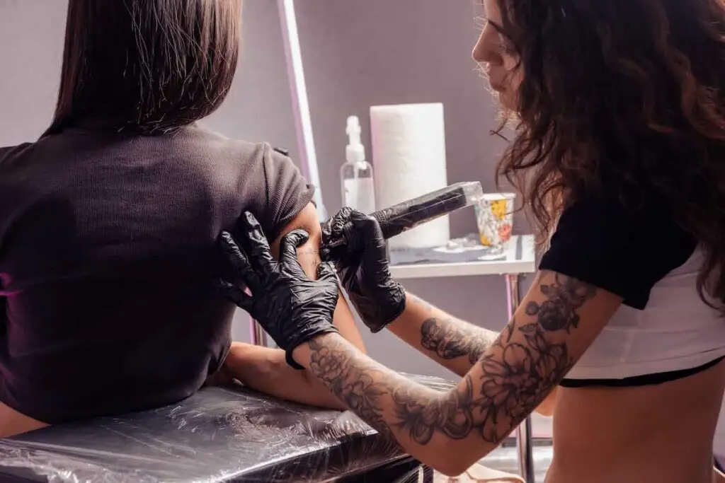 Young woman resting on an arm rest while a tattoo artist works on her upper arm.